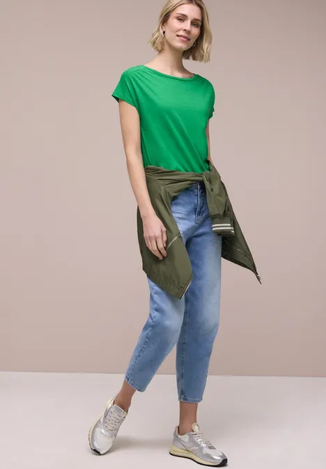 Frau trägt grünes T-Shirt, Jeans, Sneakers und hat ein Jackett um die Taille gebunden.