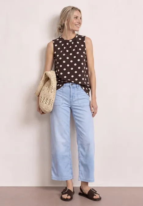 Eine Person trägt ein ärmelloses gepunktetes Top, weite hellblaue Jeans, schwarze Sandalen und eine gehäkelte Tasche.