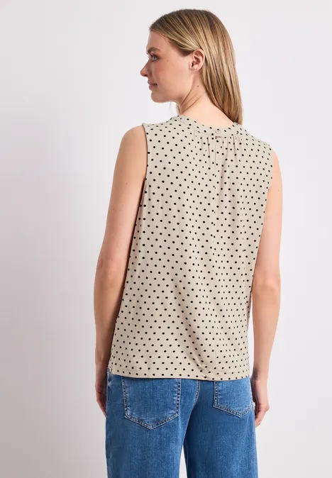 Achteraanzicht: vrouw in mouwloze beige top met zwarte stippen en spijkerbroek.