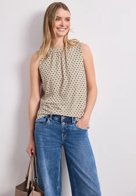 Vrouw draagt mouwloos gepatroond topje, rechte blauwe jeans en houdt een tas vast.