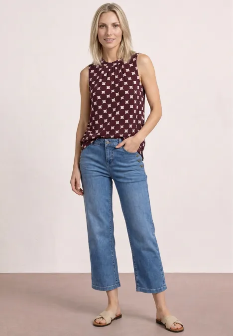 Frau vor hellem Hintergrund, trägt ein ärmelloses Top mit Muster, gerade geschnittene blaue Jeans und Sandalen.