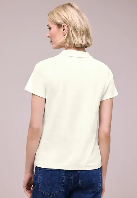 Frau in weißem Poloshirt und blauen Jeans, Rückansicht, blondes kurzes Haar, neutraler Hintergrund.