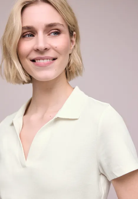 Eine Person mit kurzem blondem Haar trägt ein cremefarbenes, kurzärmeliges Poloshirt.