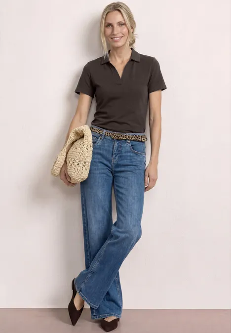 Frau in dunklem Poloshirt, ausgestellten Jeans, geflochtenem Gürtel und Strohhut.