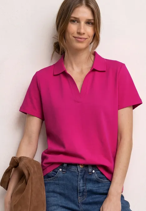 Femme portant un polo rose et un jean bleu, tenant une veste marron à la main.