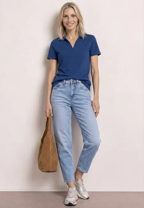 Vrouw in blauw poloshirt, lichte jeans en sneakers, met bruine tas voor lichte achtergrond.