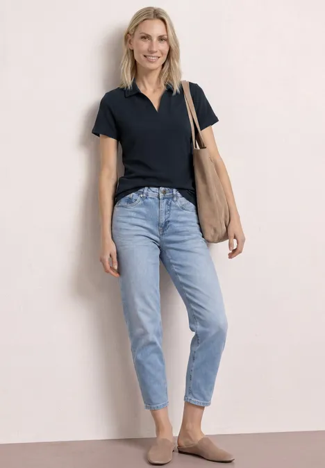 Vrouw staat voor een lichte achtergrond, draagt een zwart polohemd, blauwe jeans, bruine loafers en een schoudertas.