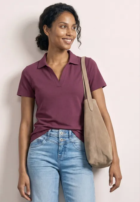 Une personne porte un polo violet, un jean bleu et un sac épaule beige devant un fond neutre.