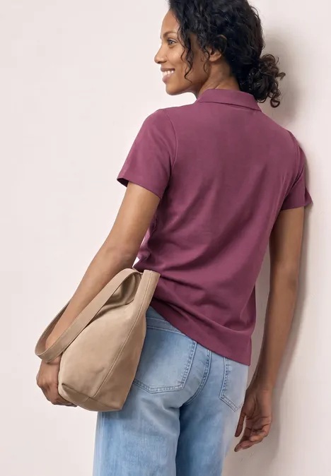 Personne de dos en polo violet et jean bleu, avec un sac beige sur l'épaule, fond uni clair.