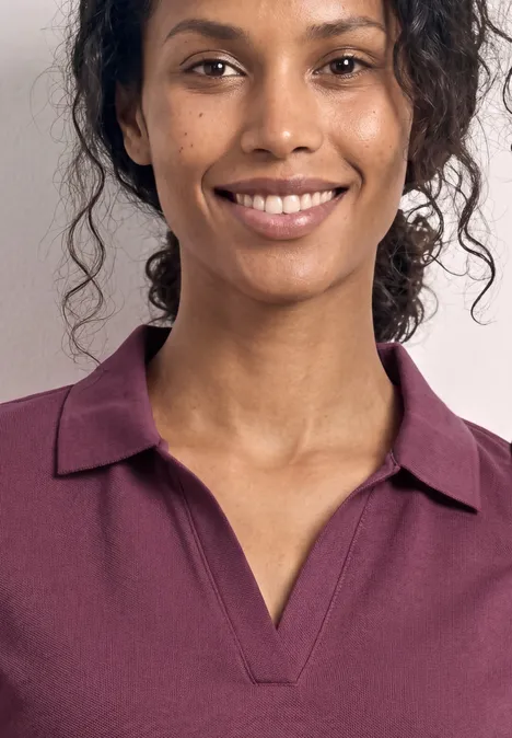 Une personne aux cheveux bouclés porte un polo violet. Seul le buste et le cou sont visibles.