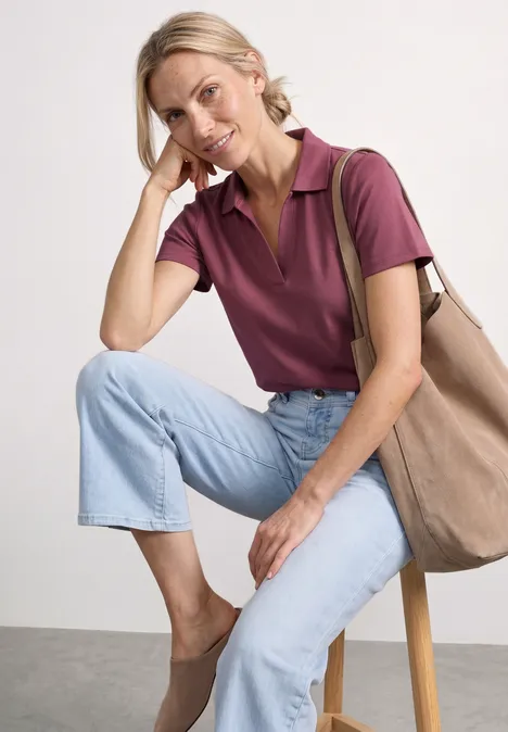 Eine Person sitzt auf einem Hocker, trägt lila Poloshirt, helle Jeans, bequeme Schuhe und eine Tasche.