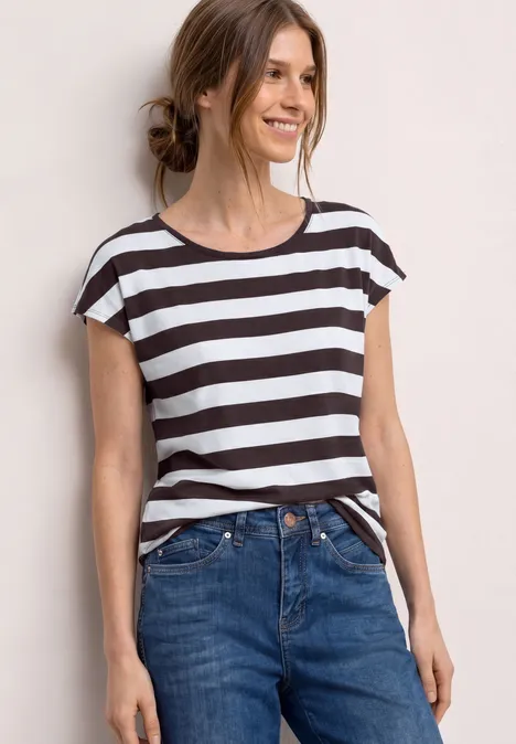 Eine Frau trägt ein gestreiftes T-Shirt und Jeans vor hellem Hintergrund.