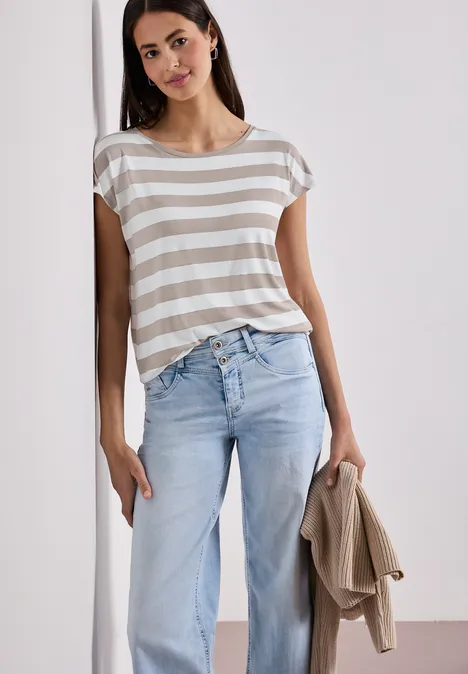 Frau lehnt an Wand, trägt gestreiftes T-Shirt, helle Jeans und hält eine beige Jacke in der Hand.