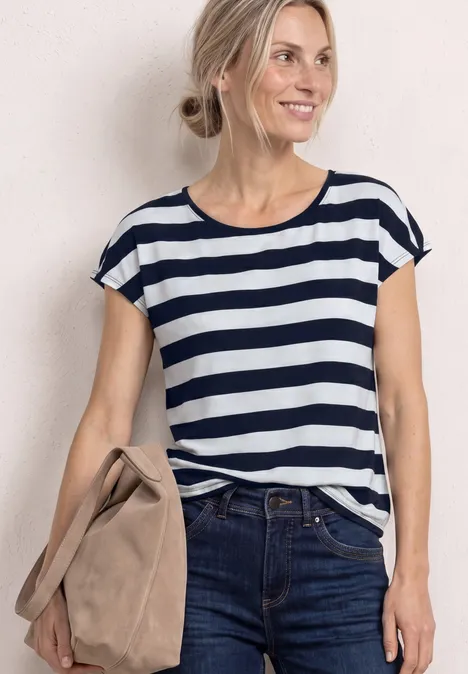 Frau in Ringelshirt hält Tasche, Jeans, vor hellem Hintergrund.