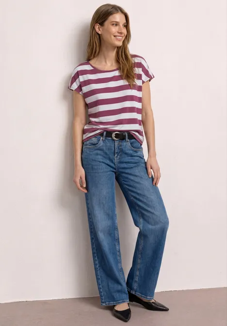 Vrouw met gestreept T-shirt, wijde spijkerbroek en platte zwarte schoenen, staand voor een lichte achtergrond.