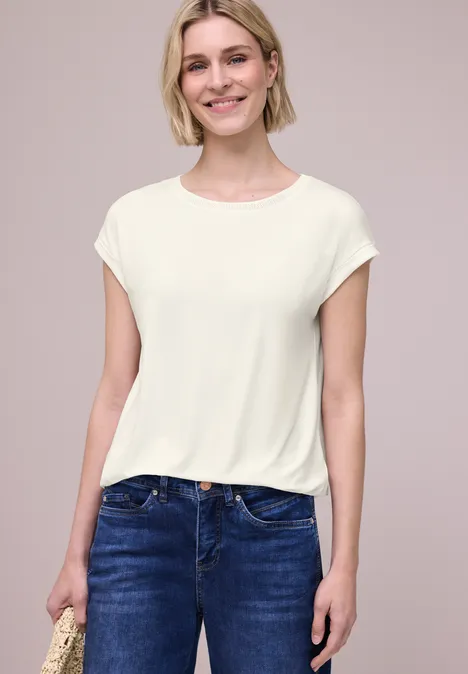 Frau trägt schlichtes weißes T-Shirt und blaue Jeans, steht vor neutralem Hintergrund.