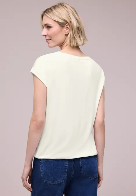 Frau mit kurzem blonden Bob trägt cremefarbenes T-Shirt und blaue Jeans, Rückenansicht vor neutralem Hintergrund.