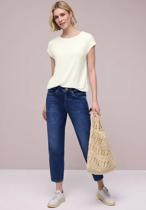 Modell trägt weiße Kurzarmshirt, blaue Jeans, weiße Sneaker und hält eine gehäkelte Strandtasche.