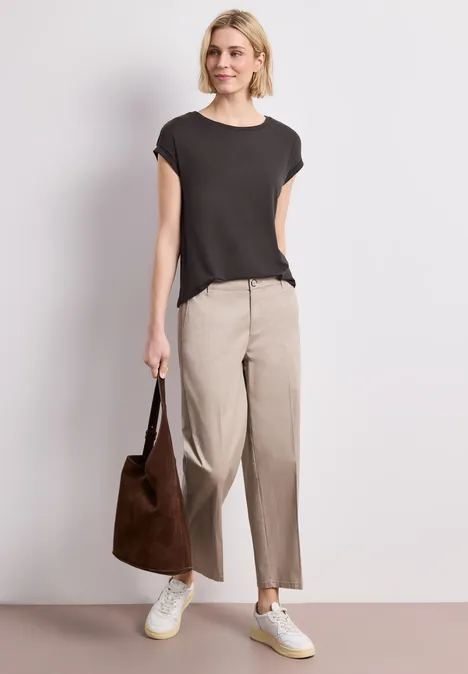 Een persoon draagt een zwart t-shirt, beige broek, witte sneakers en houdt een bruine tas vast.