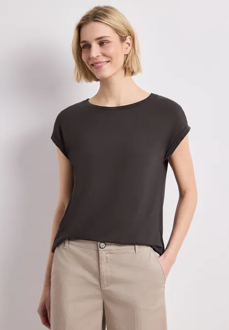 Een vrouw draagt een eenvoudig zwart t-shirt en een beige broek, poseert met één hand in de zak tegen een neutrale achtergrond.