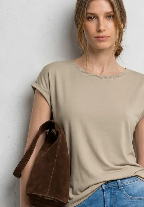 Femme en t-shirt beige et jean bleu, tenant un sac en daim marron sur l'épaule devant un mur clair.