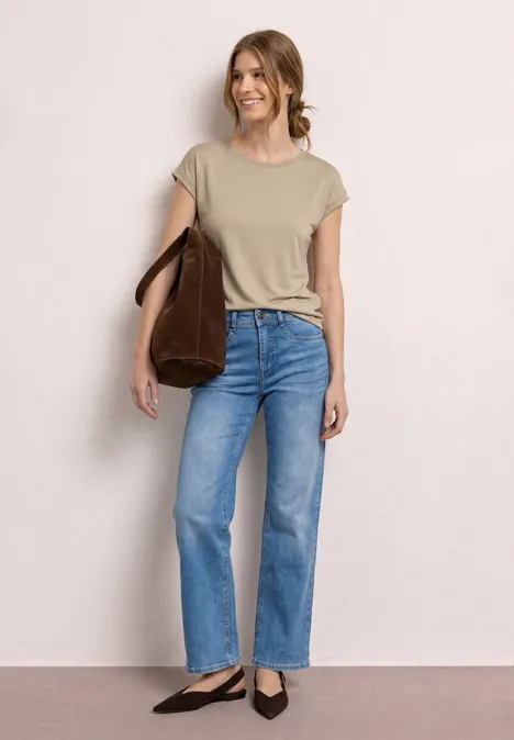 Une femme porte un t-shirt beige, un jean bleu, des chaussures noires et un sac marron devant un fond neutre.