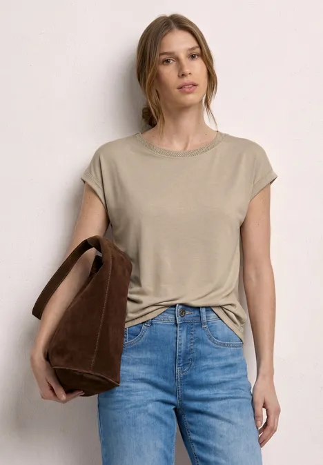 Femme en t-shirt beige à manches courtes et jeans bleus, portant un sac marron devant un mur neutre.