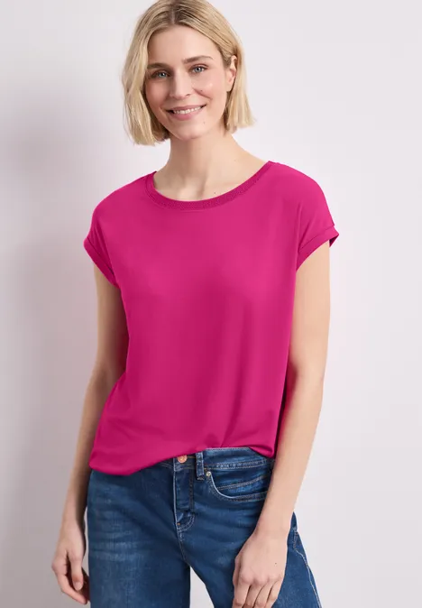 Modell trägt pinkes T-Shirt und blaue Jeans vor hellem Hintergrund.