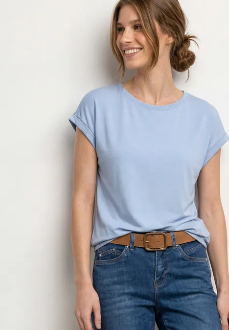 Une femme porte un t-shirt bleu clair à manches retroussées, un jean et une ceinture marron.