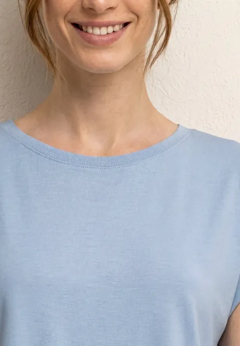 Une femme souriante en t-shirt bleu clair devant un mur neutre.