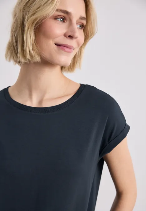 Vrouw in zwart T-shirt met opgerolde mouwen, licht zijwaarts gekeerd, glimlacht.