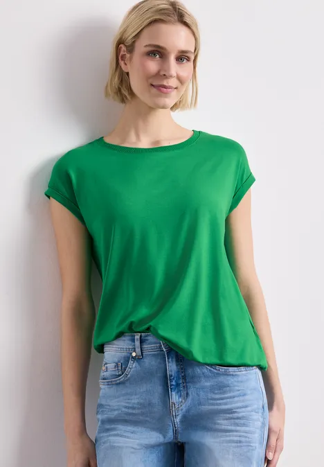 Frau in grünem T-Shirt und blauer Jeans vor weißem Hintergrund.