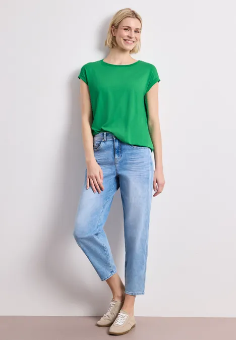 Ein Modell steht vor weißem Hintergrund in grünem T-Shirt, hellblauen Jeans und beigen Sneakern.