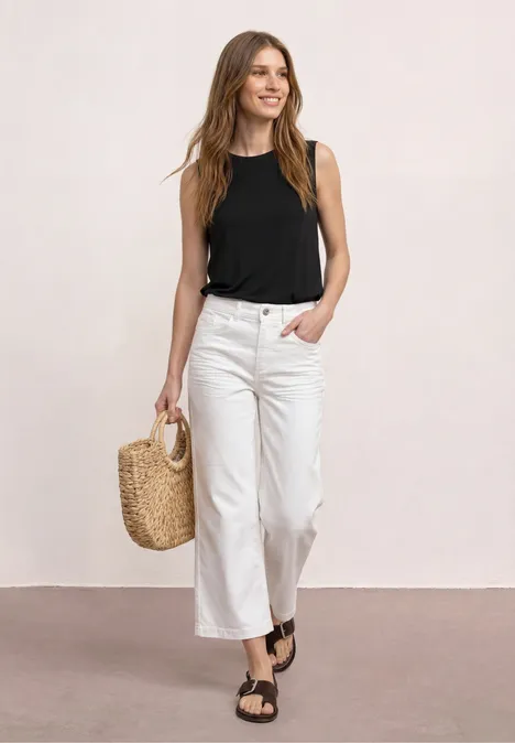 Frau in weißer Culotte, schwarzem Top, leichter Strohtasche und schwarzen Sandalen vor hellem Hintergrund.