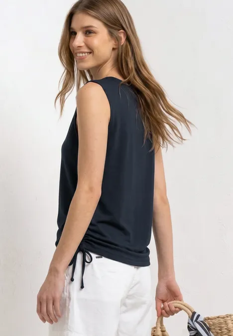 Vrouw in een zwarte mouwloze top, witte broek en strooien tas, ontspannen staand op de foto.