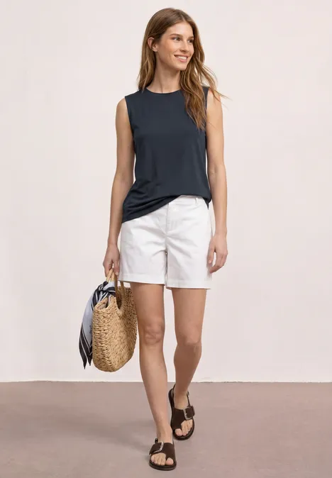 Vrouw in marineblauw topje en witte shorts, met een gevlochten tas en zwarte sandalen voor een neutrale achtergrond.