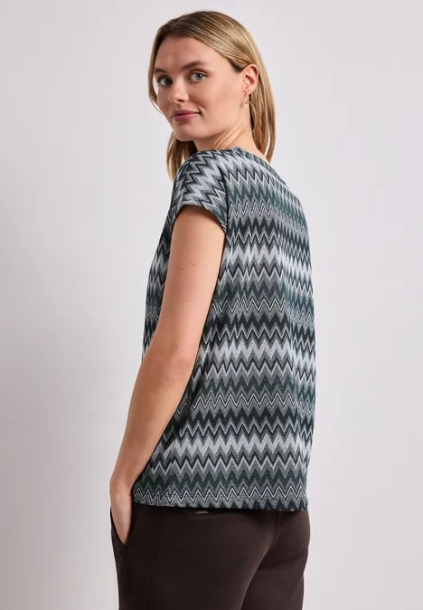 Vrouw van opzij in een grijs-groen-zwart zigzagtop met korte mouwen en een donkere broek.