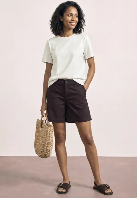 Frau trägt weißes T-Shirt, schwarze Shorts, schwarze Sandalen und hält eine geflochtene Tasche.