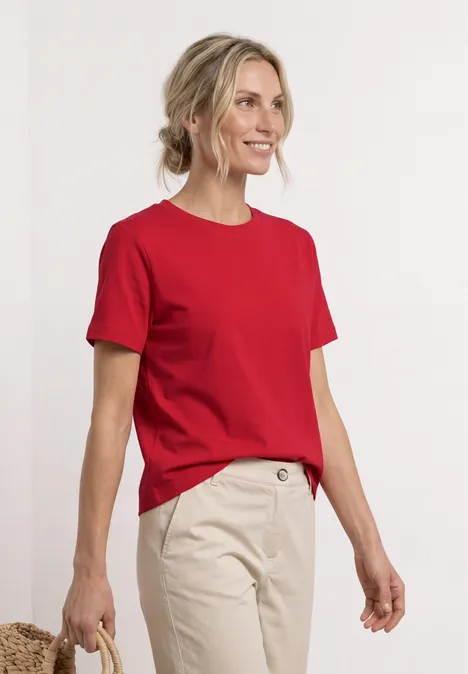 Person in rotem T-Shirt und beiger Hose steht vor weißem Hintergrund, Gesicht unscharf.