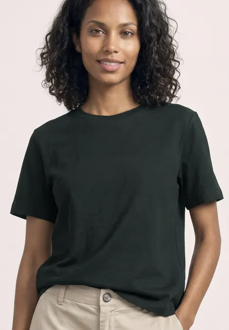 Eine Frau mit lockigem Haar trägt ein schlichtes schwarzes T-Shirt und helle Hosen.