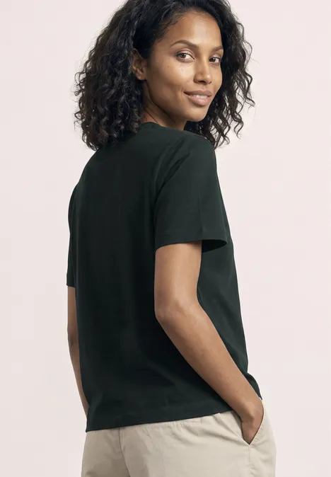 Eine von hinten abgebildete Frau in schwarzem T-Shirt und beigen Hosen, die mit den Händen in den Taschen steht.