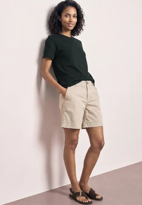 Eine Person lehnt sich an eine Wand, trägt ein schwarzes T-Shirt, beige Shorts und schwarze Sandalen.