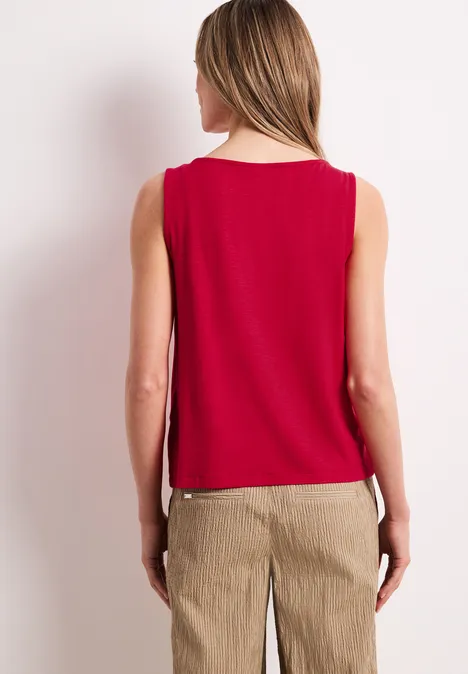 Frau von hinten in rotem ärmellosen Top und beigen Hosen vor neutralem Hintergrund.