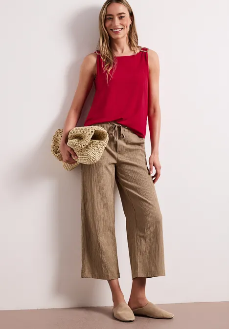 Frau in rotem ärmellosem Top, beigen Culotte-Hosen und Slippern, mit Strohtasche vor hellem Hintergrund.