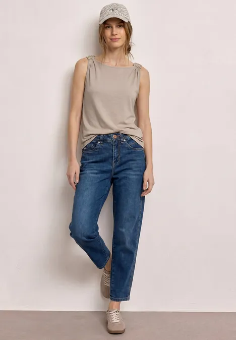 Une personne pose devant un mur clair, vêtue d’un débardeur beige noué, d’un jean bleu et de mocassins assortis.