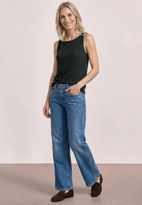 Frau trägt schwarzen ärmellosen Top und blaue Jeans, steht locker mit Händen in den Taschen.