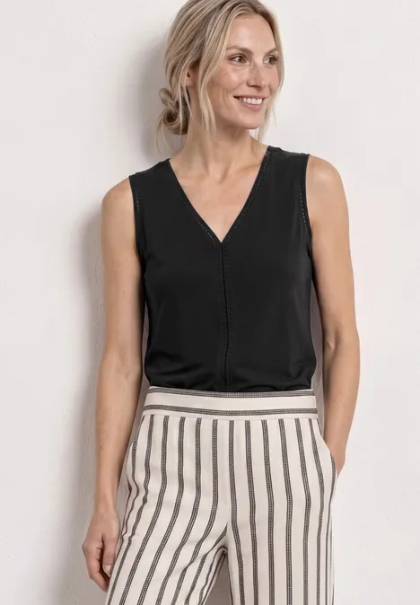 Vrouw in een zwart mouwloos topje en beige broek met zwarte strepen, handen in de zakken.