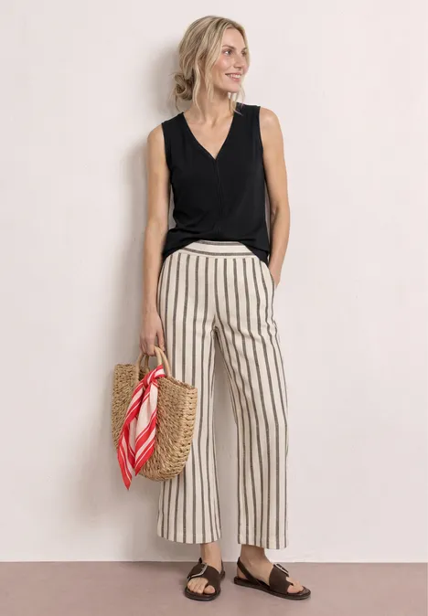 Vrouw in zwart hemd, gestreepte broek, bruine sandalen en rieten tas met rode sjaal tegen lichte achtergrond.