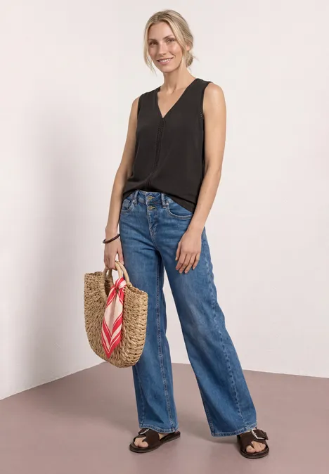 Frau in schwarzem ärmellosen Top, weiten Jeans und Sandalen trägt eine Strohtasche mit rotem Schal.