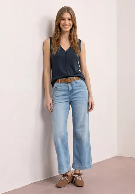 Eine Frau trägt ein dunkelblaues Top, helle weite Jeans und braune Schuhe vor einer hellen Wand.